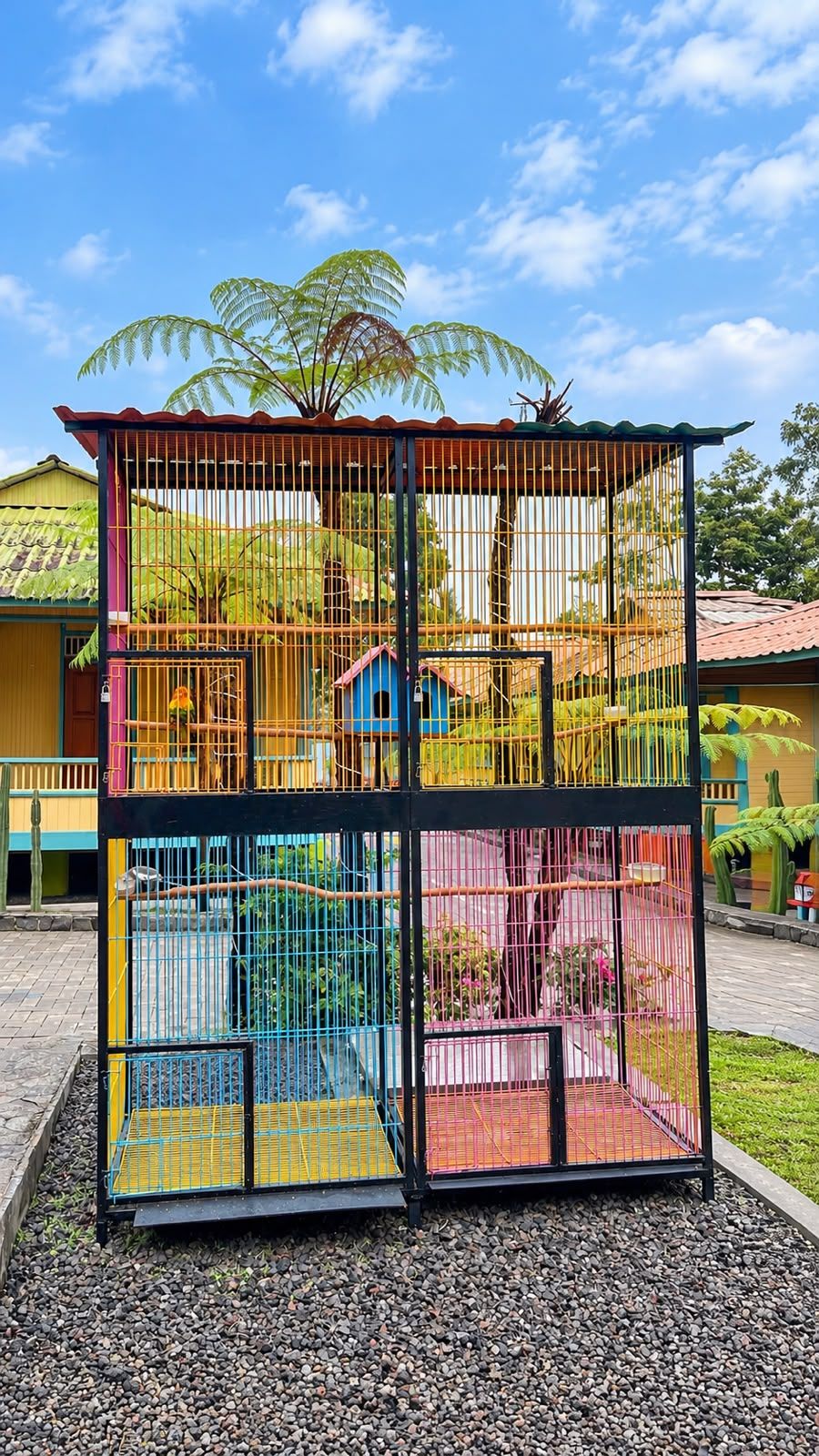 A multi-level birdcage structure with various compartments, set against a backdrop of a clear sky and tropical vegetation.