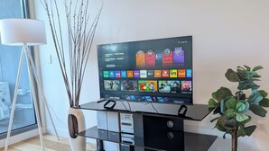 Una sala de estar moderna con un televisor de pantalla plana sobre un soporte negro, un jarrón blanco con ramas, una planta en maceta y una lámpara de pie.