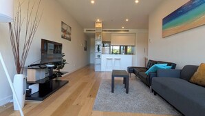 Una sala moderna con un sofá, una mesa de centro y un televisor sobre un mueble bajo. Se ve una zona de cocina con una barra y taburetes al fondo.