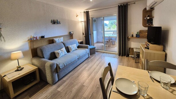 Ein Wohnzimmer mit einer Couch, einem Esstisch mit Stühlen, einem Fernseher und einer Glastür, die auf einen Balkon führt.