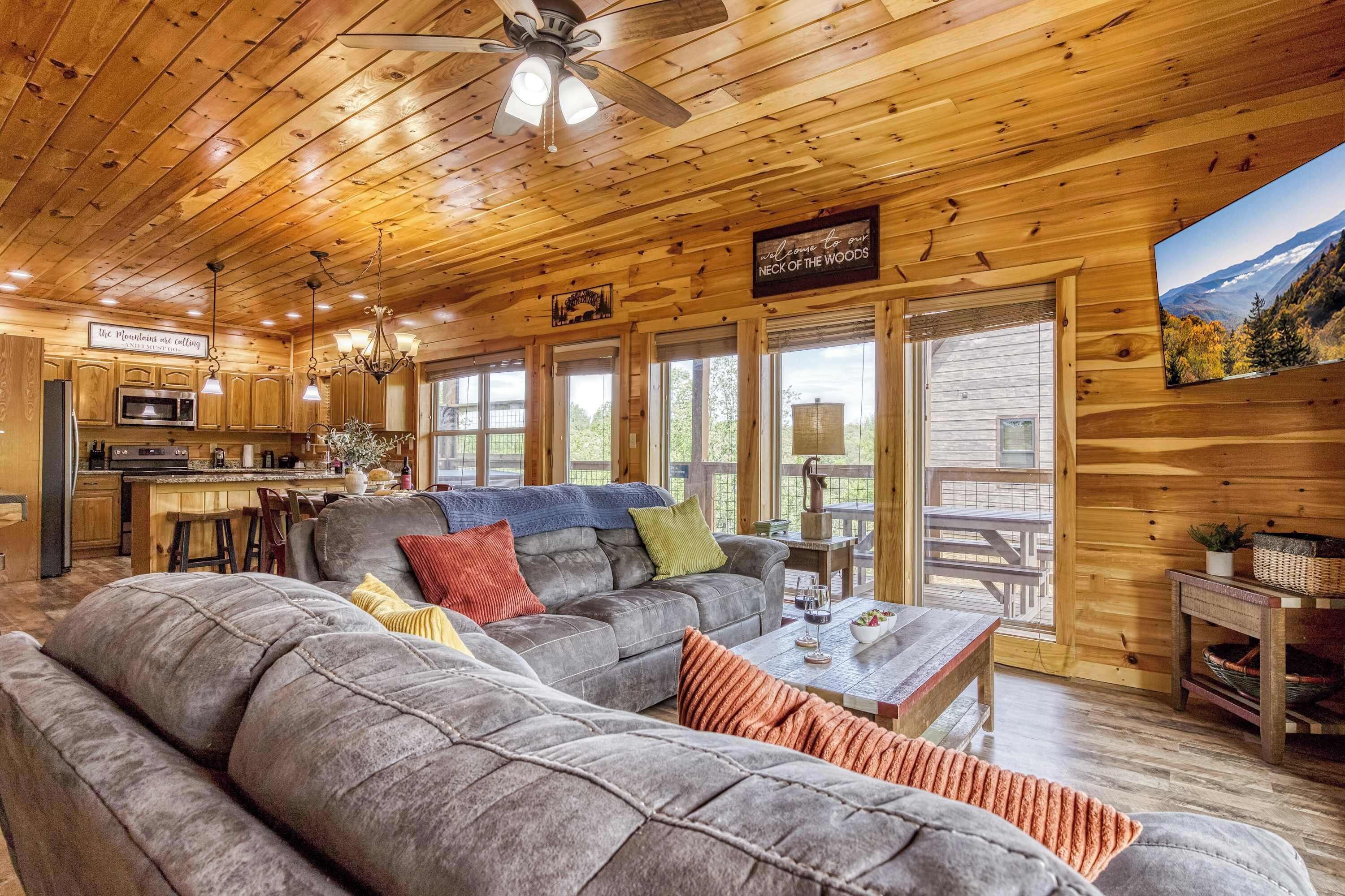 A wooden cabin interior with a large sofa, dining table, kitchen area, and a scenic view through the windows.