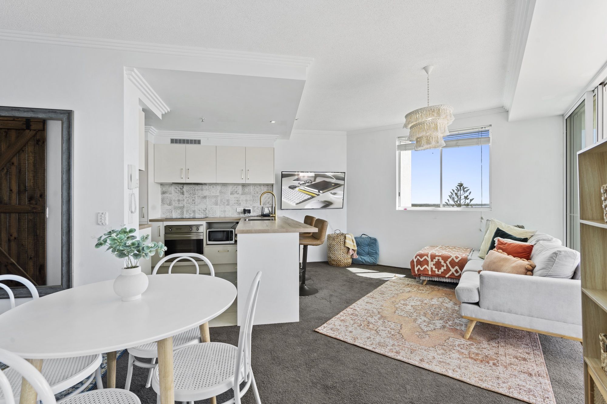 Een moderne keuken met witte kasten, een eethoek met een witte tafel en stoelen, en een woonkamer met een bank en een kamerbreed vloerkleed.