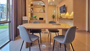 A modern dining area with a large table, four grey chairs, pendant lights, a flat-screen TV, and a view of the outdoors. - LeGacy By SanLiving - 3BR - PIK2 (Kosambi)