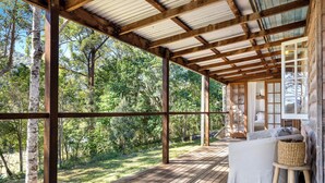 A wooden deck with a covered area, a white sofa, and a wicker stool. - Conondale Lodge – Peaceful Country Escape (Conondale)