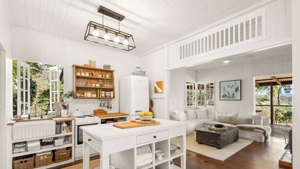 A spacious living area with a kitchen, white walls, wooden flooring, and large windows offering views of the outdoors. - Conondale Lodge – Peaceful Country Escape (Conondale)