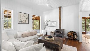 A cozy living room with a white couch, a wooden coffee table, a fireplace, and large windows with a view of trees. - Conondale Lodge – Peaceful Country Escape (Conondale)