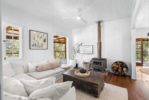 Ein gemütliches Wohnzimmer mit einem weißen Sofa, einem Holztisch, einem Kamin und großen Fenstern mit Blick auf Bäume.