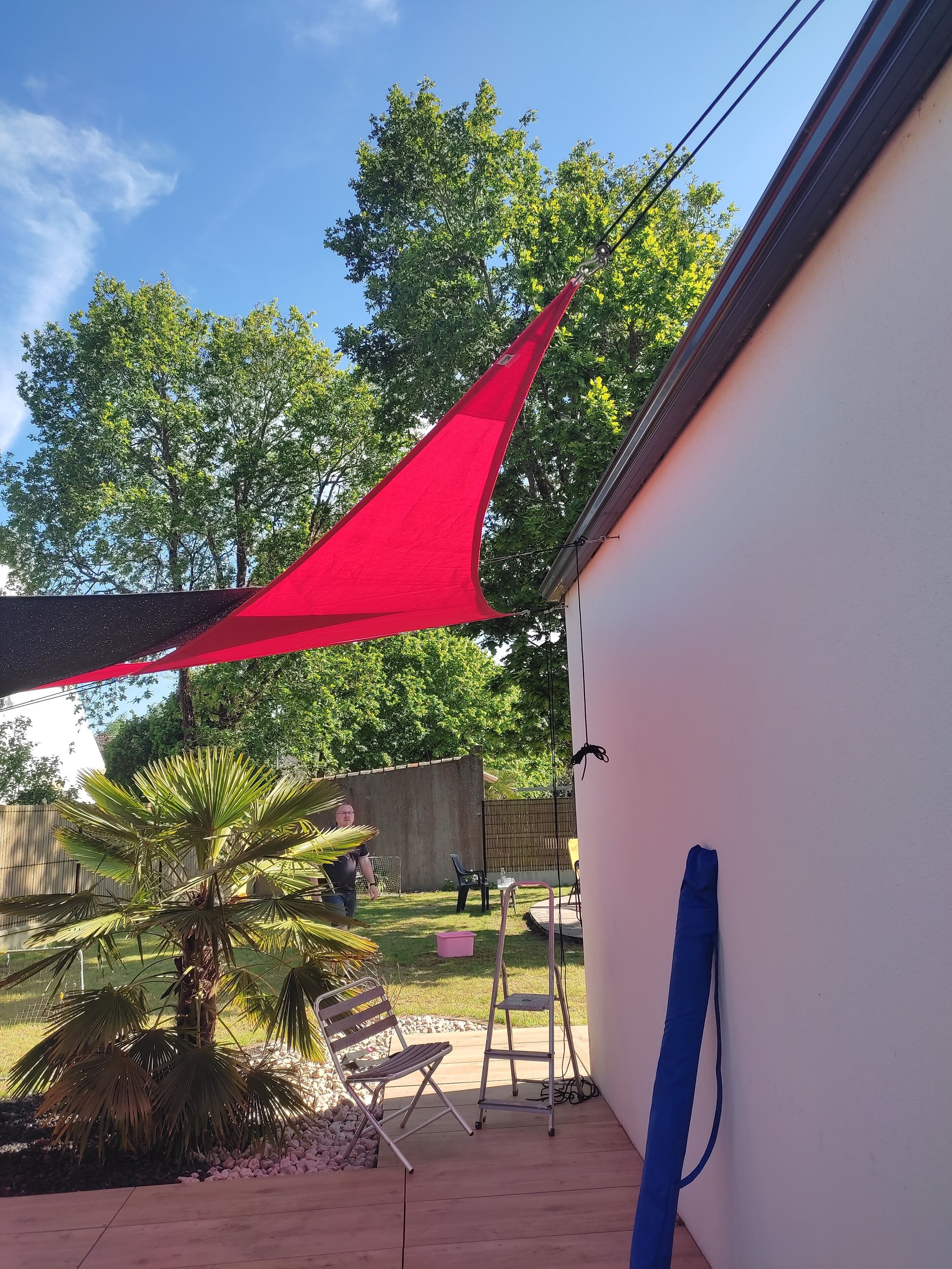 A patio area with a red shade sail, a palm tree, outdoor furniture, and a ladder.