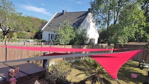 Ein Garten mit einer Holzterrasse, einem roten Sonnensegel und einem Haus im Hintergrund.