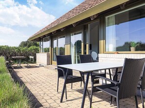 Een terras buiten met een tafel en stoelen, een geplaveid pad en een gebouw met grote ramen en een pannendak.