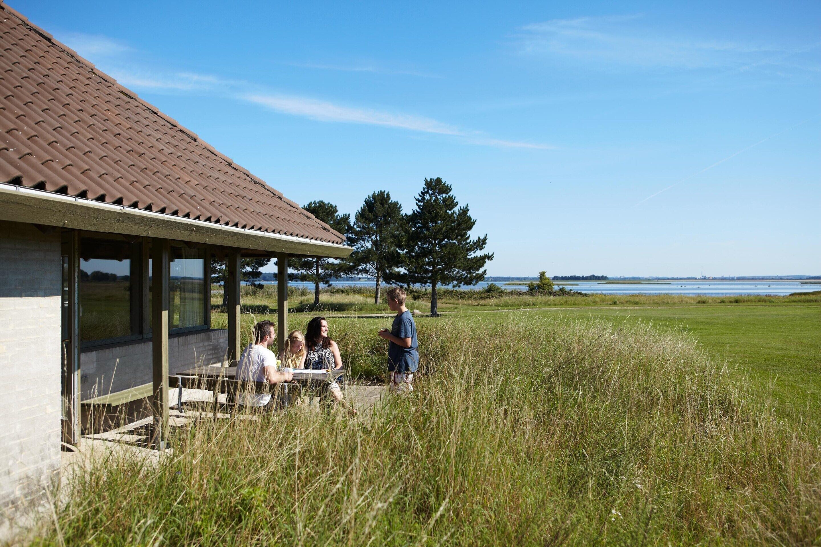 A building with a tiled roof and large windows, surrounded by tall grass and a view of a body of water.