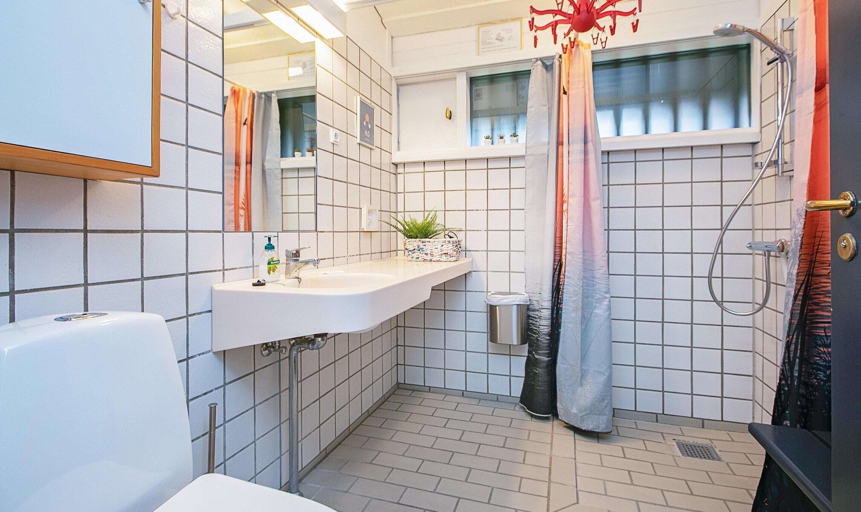 Une salle de bain avec un lavabo blanc, un miroir et une douche. On peut voir une plante en pot sur le lavabo et une poubelle à côté.