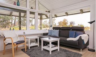 Una sala de estar acogedora con un sofá, un sillón, una mesita y una lámpara de pie. Amplias ventanas ofrecen vistas al exterior.