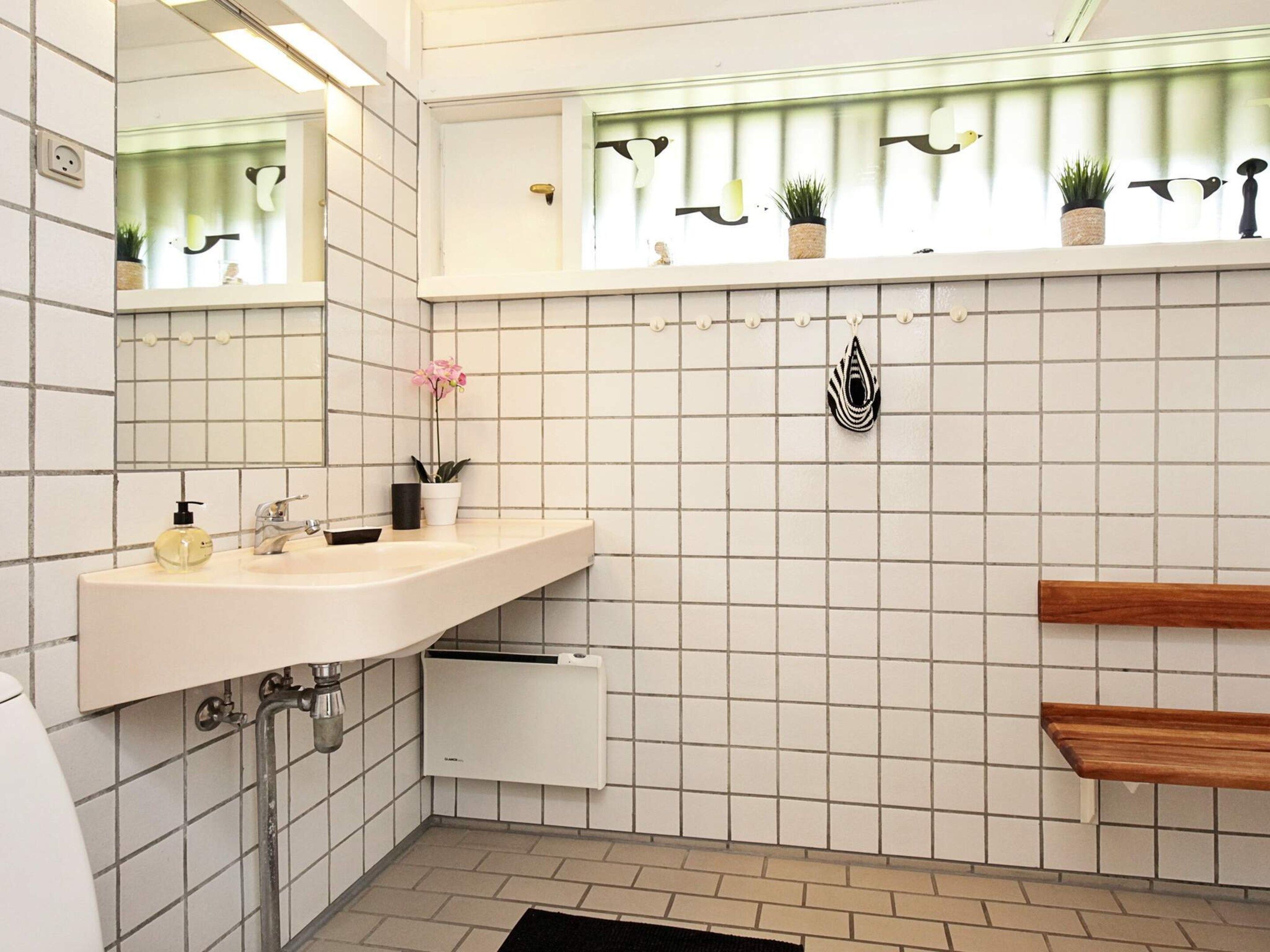 A bathroom with a white tiled wall, a mirror, a sink with a soap dispenser, a potted plant, and a shelf with decorative items.