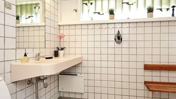 A bathroom with a white tiled wall, a mirror, a sink with a soap dispenser, a potted plant, and a shelf with decorative items.