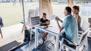 A covered outdoor seating area with a table and chairs, a person playing cards, and a view of tents and a car in the background. - Begur Nature Glamping for 5 Guests (Begur)