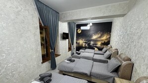 Ein Zimmer mit einem großen Bett, einem kleineren Bett und einer Schlafcouch. Es gibt ein Fenster mit Vorhängen, einen wandmontierten Fernseher und ein Wandgemälde mit einer Nachtszene.