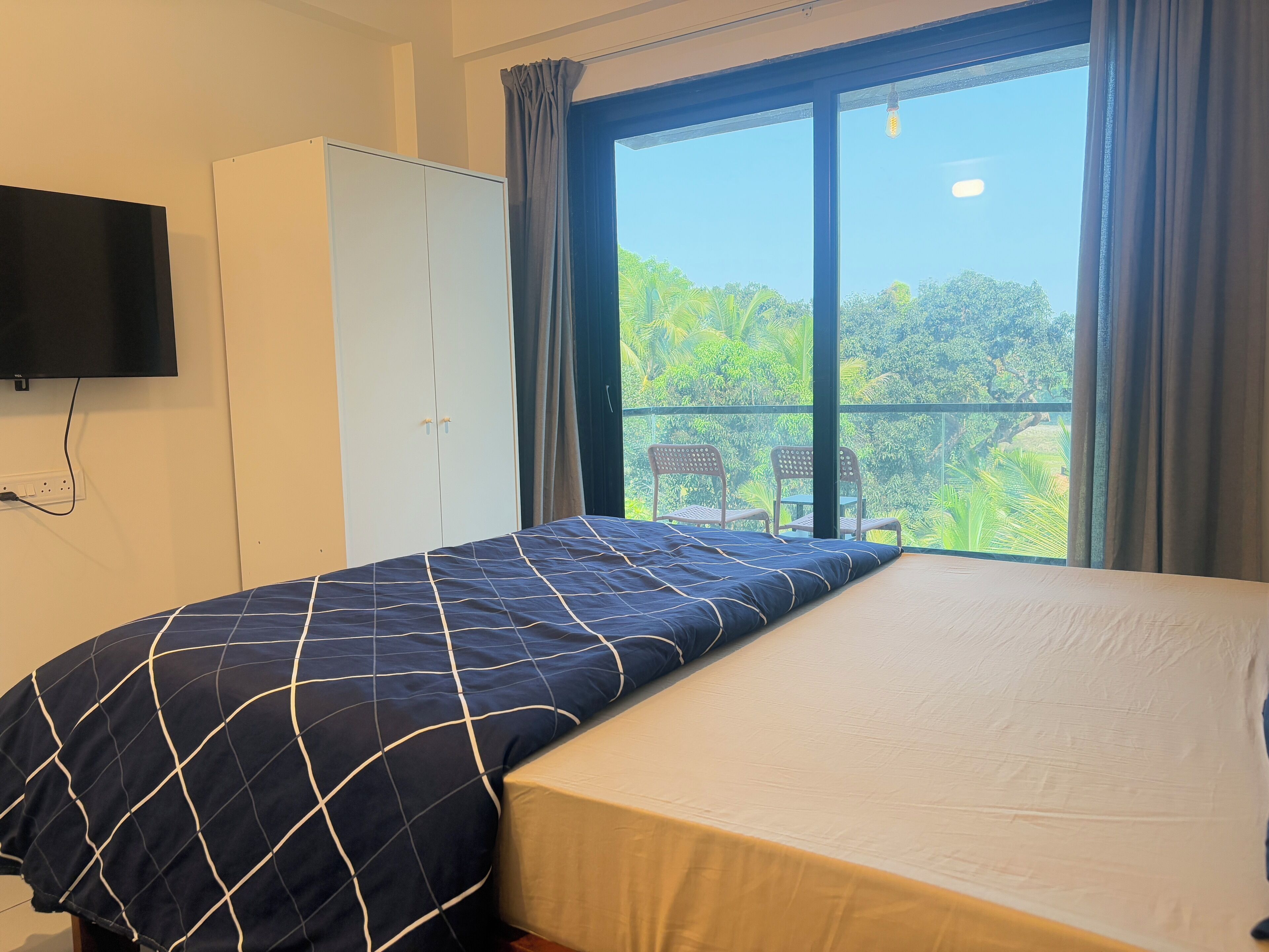 A room with a bed, a wardrobe, a television, and a window with a view of greenery.