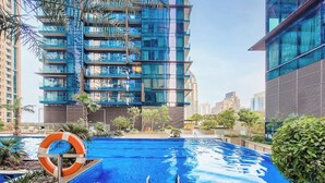 A modern building with a glass facade, a swimming pool with a lifebuoy, and surrounding greenery. - The Residence at Marina Gate 2  (Dubai)