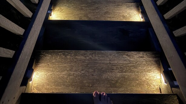 Un escalier en bois avec des rampes, qui monte, et les pieds d’une personne en bas.