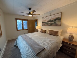 Une chambre à coucher avec un lit, un ventilateur de plafond, une fenêtre, un tableau, une table de chevet et une lampe.