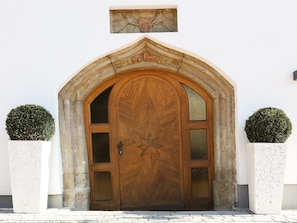 A wooden arched door with a geometric pattern, flanked by two potted topiaries, and a stone plaque above. - Composite penthouse with shared wellness (Piesendorf)