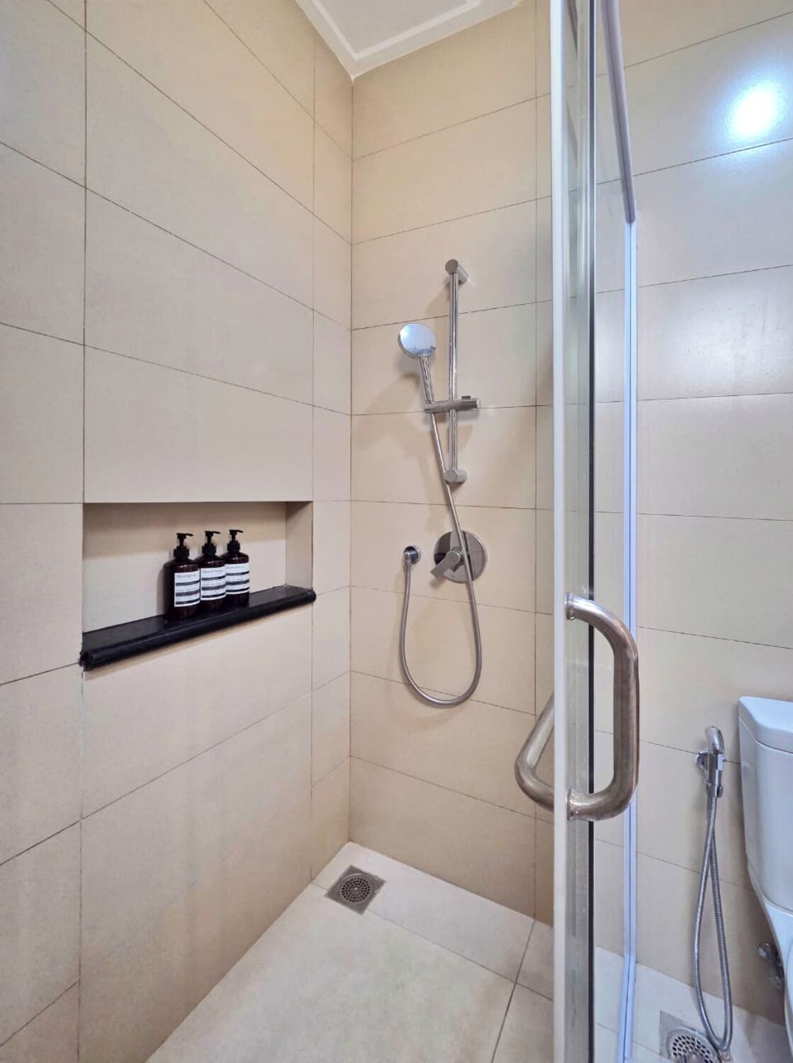 A small, modern bathroom with a glass shower enclosure, a black shelf with toiletries, and a toilet visible to the right.