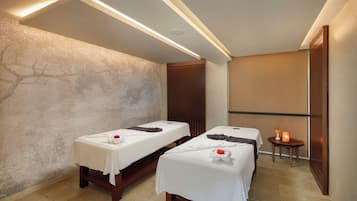 Un cuarto de spa con dos camillas de masaje, un revestimiento de pared de madera y una mesita con piedras calientes.