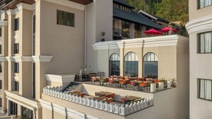 Een hotel met meerdere verdiepingen, buiten zitgedeeltes, een balkon met rode parasols en uitzicht op een bergachtig landschap.