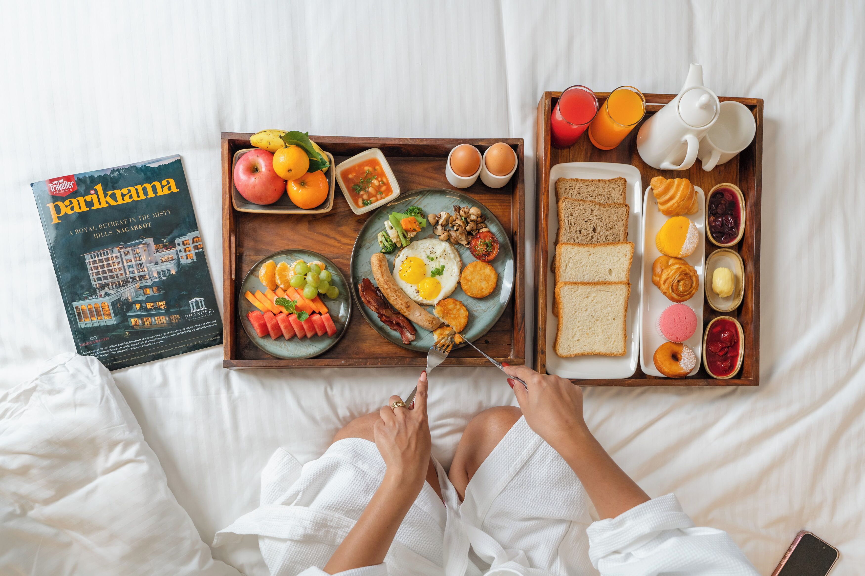 A breakfast tray with eggs, bacon, sausages, toast, and pastries on a bed.