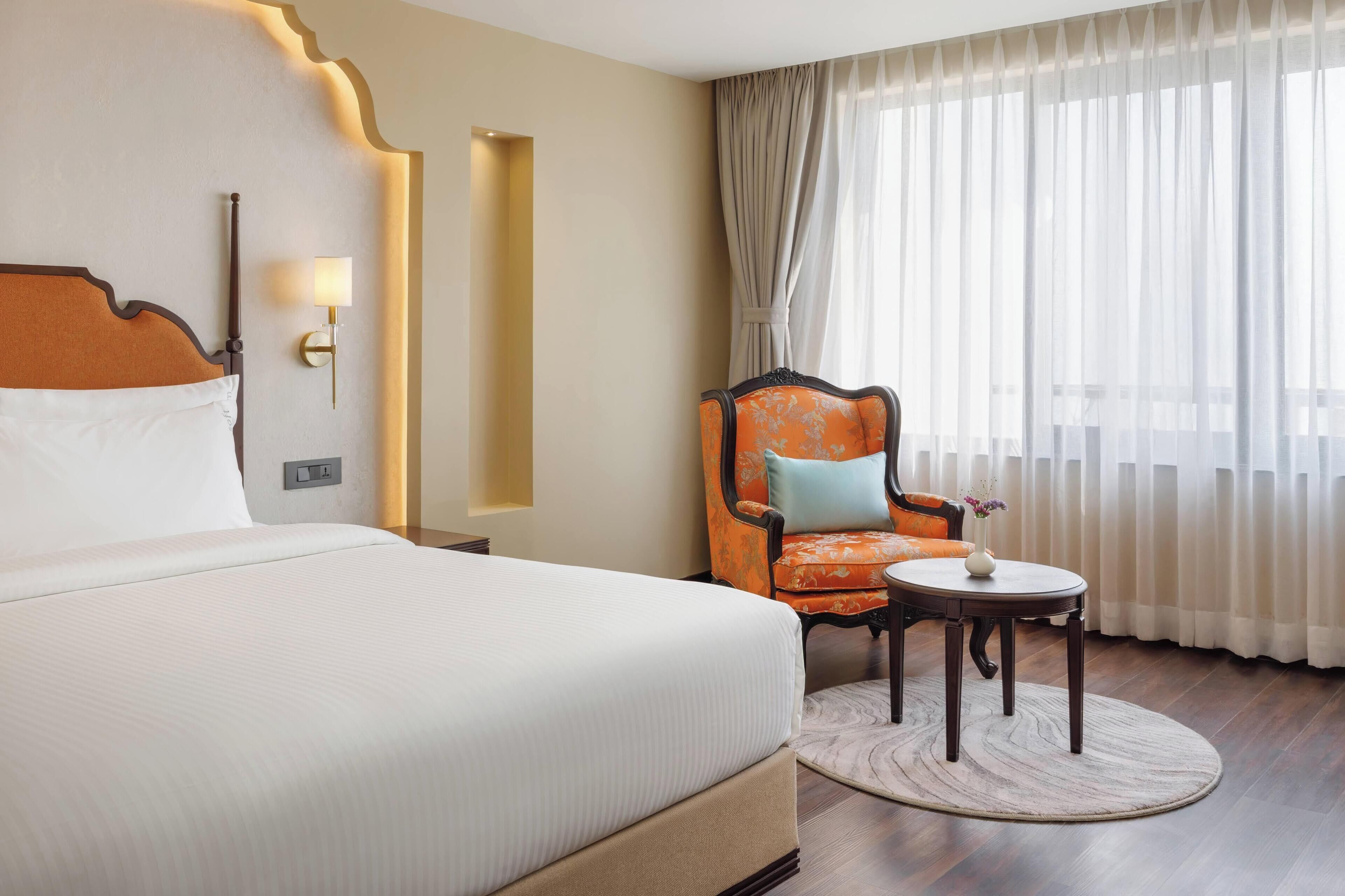A hotel room with a large bed, a chair, a small table, and a window with sheer curtains.