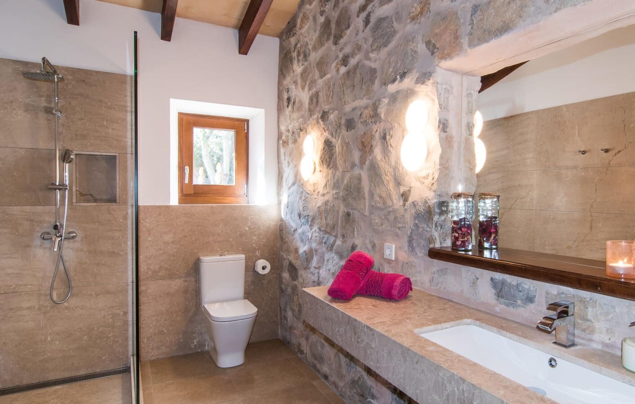 A bathroom with a stone wall, a shower, a toilet, a sink, and a window.
