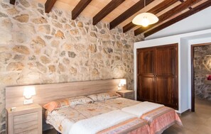 A bedroom with a stone wall, wooden ceiling beams, a double bed with floral bedding, bedside lamps, and a wooden wardrobe. - Stunning home in Arta with outdoor swimming pool (Arta)