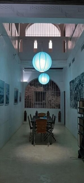 Un espace repas doté d’un plafond haut, de deux lanternes bleues suspendues, d’une table avec des chaises et d’une grande fenêtre avec des vitres à croisillons.