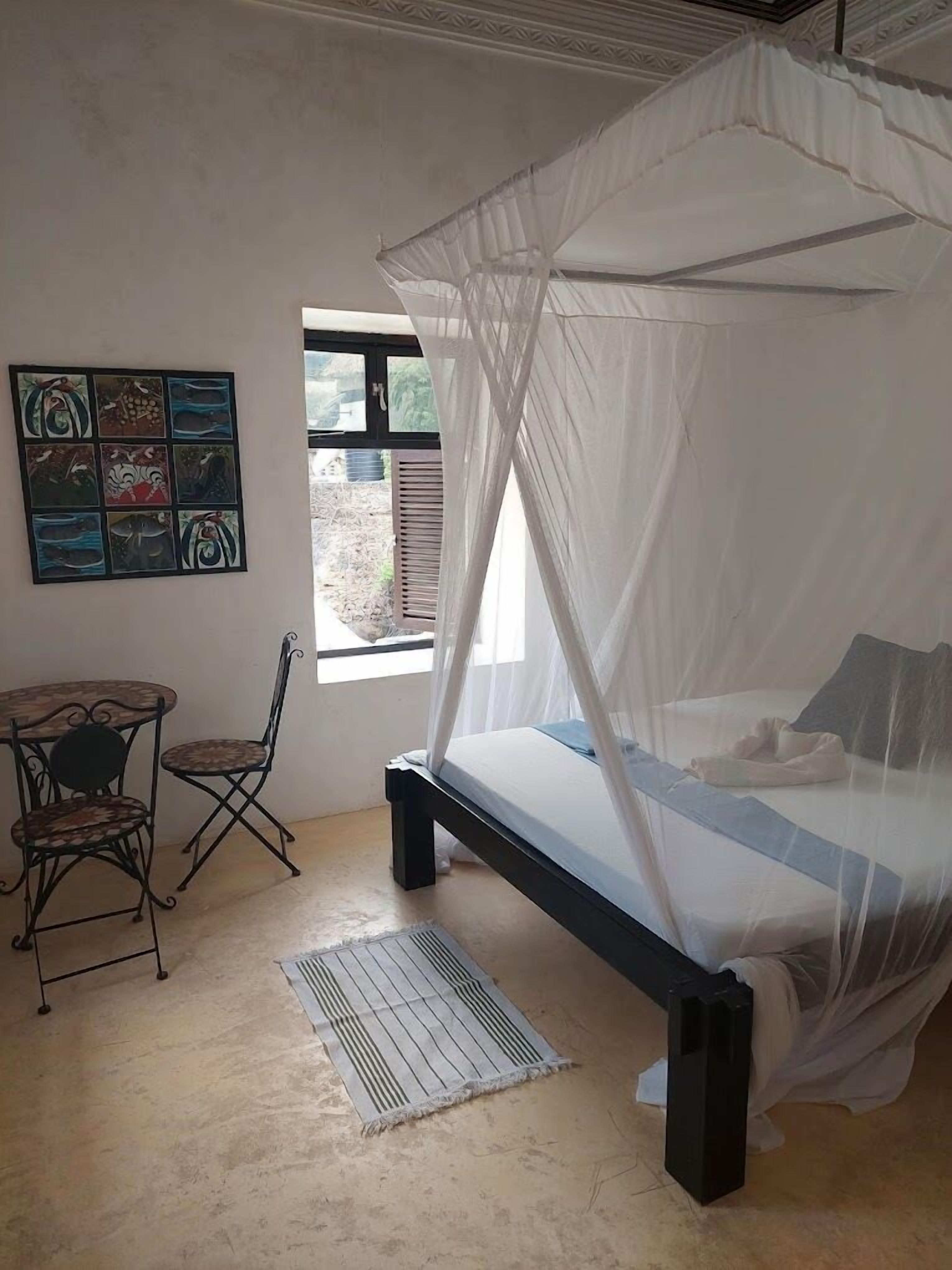 Een slaapkamer met een bed onder een baldakijn, een klein tafeltje met stoelen en een raam met uitzicht.