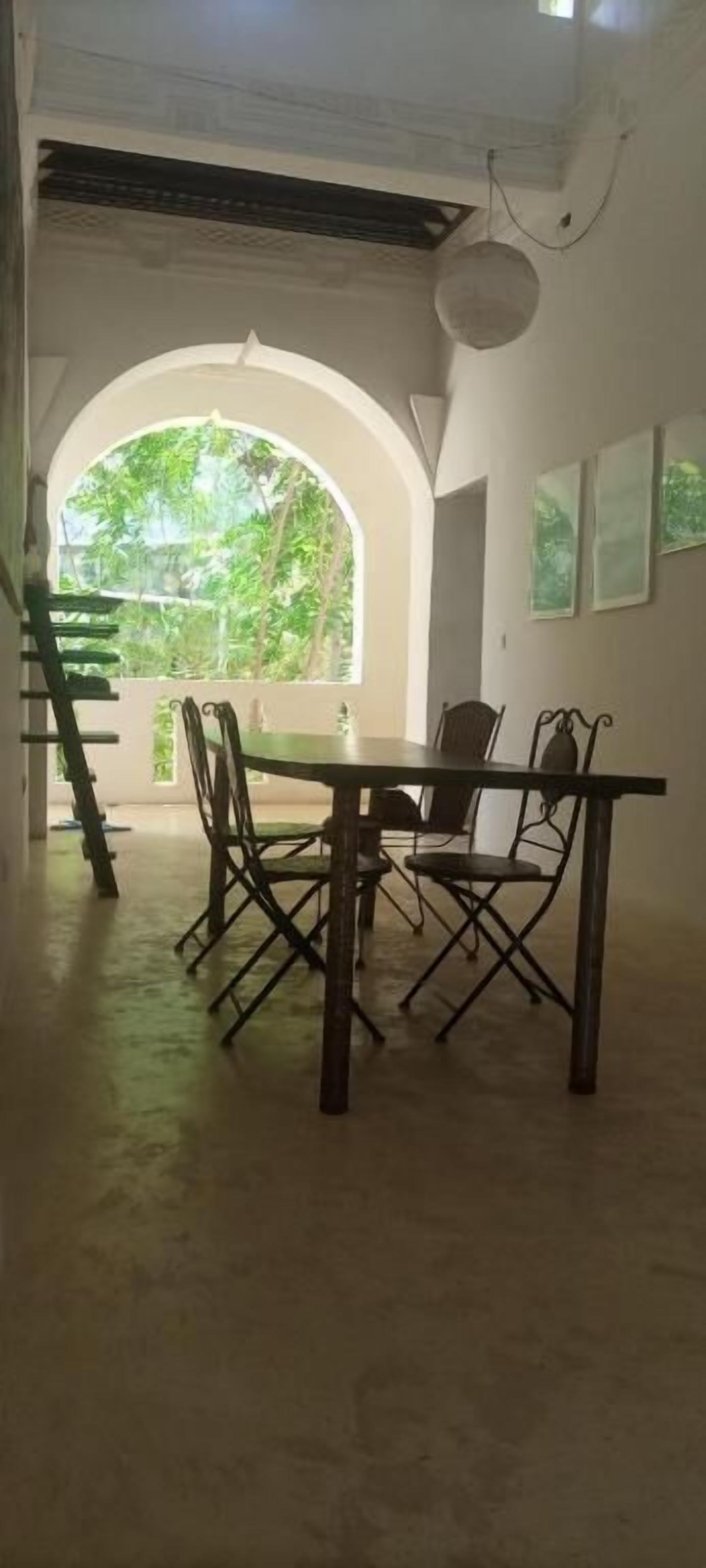 Un coin repas avec une table et des chaises, une fenêtre en arc de cercle donnant sur un paysage verdoyant et un ventilateur de plafond.