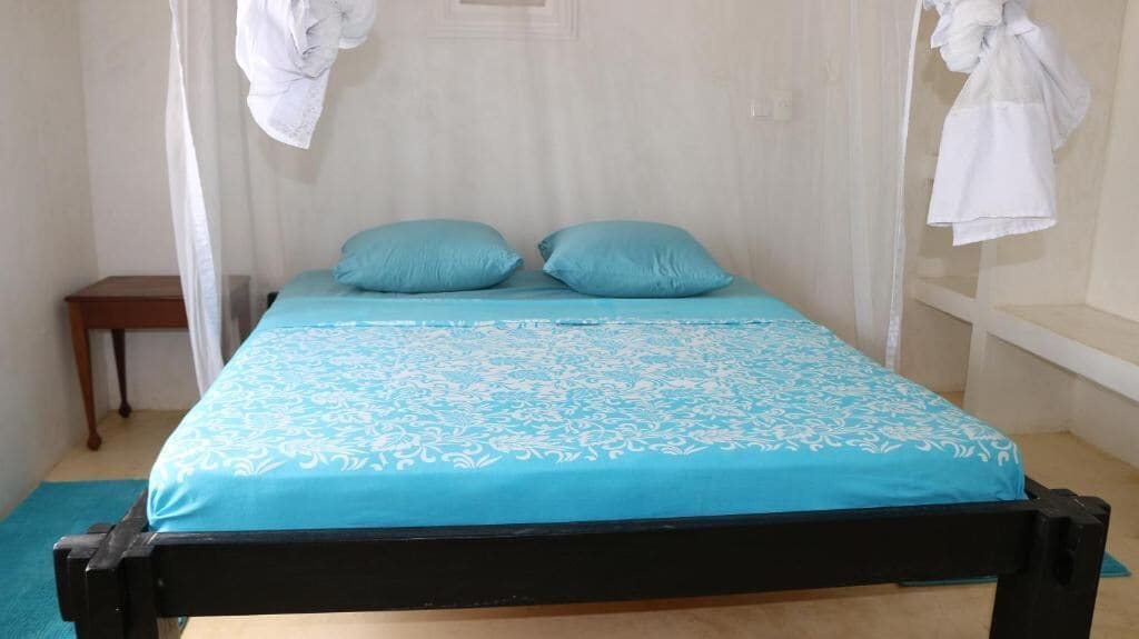 Ein Bett mit blauer Bettdecke und zwei Kissen, ein Nachttisch aus Holz und ein weißer Vorhang.