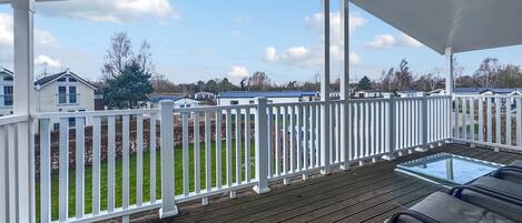 A covered outdoor area with a wooden deck and white railings, overlooking a grassy area with houses in the background.