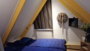 Quarto sótão pequeno e triangular com teto inclinado, uma cama de solteiro com roupa de cama azul, um radiador, uma TV fixada na parede e uma pequena mesa de cabeceira de madeira.