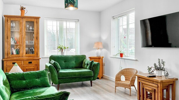 A living room with a green sofa, a wooden cabinet, a wicker chair, a small table, and a TV mounted on the wall.