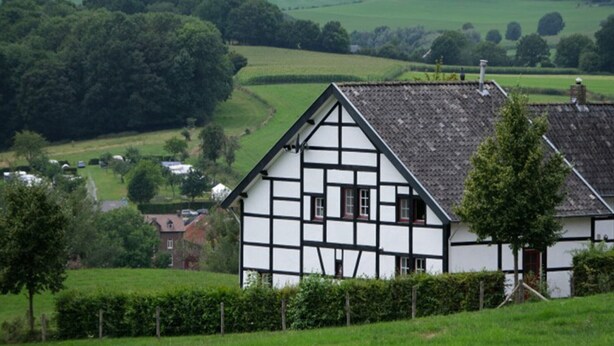 Ein traditionelles Fachwerkhäuschen mit steilem Dach, umgeben von üppigen grünen Feldern und einem dichten Wald im Hintergrund.