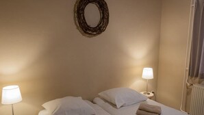 Ein Hotelzimmer mit zwei Betten, jedes mit einem weißen Kopfkissen und einem gefalteten Handtuch. Auf runden Tischen stehen je zwei Nachttischlampen. Über den Betten hängt eine runde Wanddekoration.