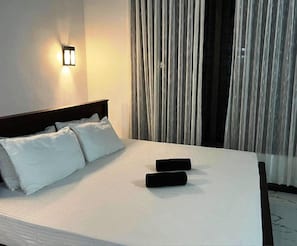 Un lit bien fait, avec du linge de lit blanc et deux oreillers noirs, une lampe fixée au mur et une vue sur un immeuble à travers des rideaux transparents.