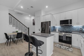 A modern kitchen with white cabinets, a stainless steel refrigerator, a built-in microwave, and a gas stove. There is a dining area with a table and chairs, and a staircase leading to another level. - LUX Houston 3BR Close To TMC, NRG, Downtown (Houston)