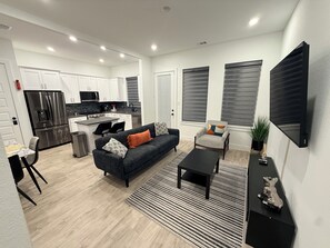 Ein stilvolles Wohnzimmer mit einer Couch, Couchtisch und einer Küchenzeile mit weißen Schränken und einem Edelstahlkühlschrank.