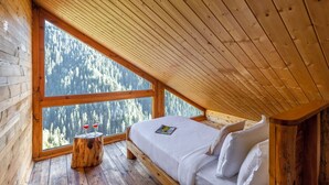 Quarto de cabana de madeira com uma janela grande, uma cama e um pequeno banquinho de madeira.