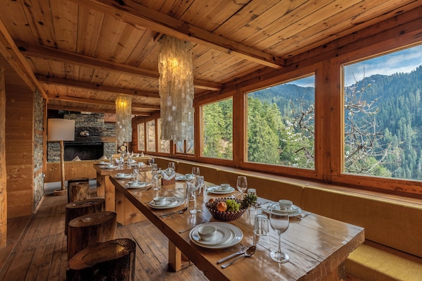 A wooden dining table set with plates, glasses, and a fruit basket, with a mountain view through large windows.