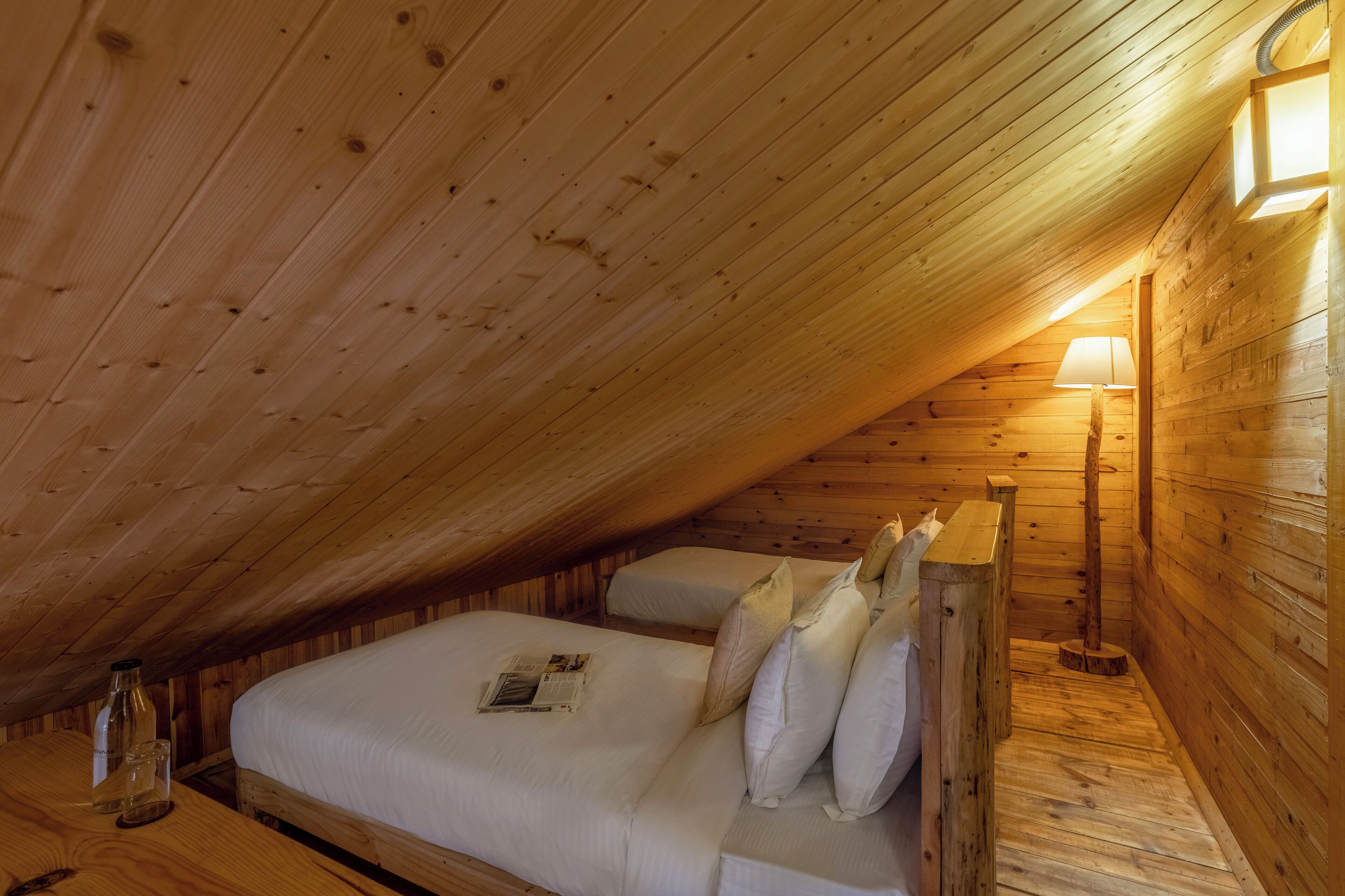 A cozy attic bedroom with wooden walls and a sloped ceiling. There is a bed with pillows and a bedside table with a lamp.