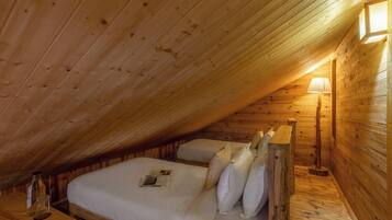 A cozy attic bedroom with wooden walls and a sloped ceiling. There is a bed with pillows and a bedside table with a lamp.