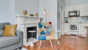 Un soggiorno con camino, un divano, una poltrona colorata e un angolo cucina con mobili ed elettrodomestici bianchi.